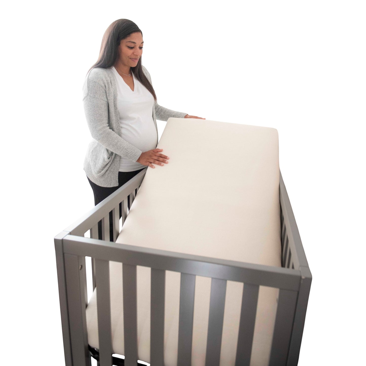 A pregnant woman placing a Modugreen Baby breathable organic 2-stage crib mattress into a grey wooden crib.