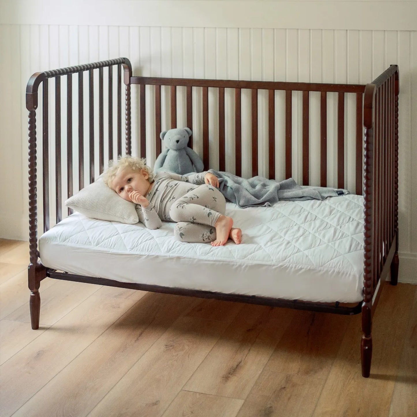 Modugreen Baby organic quilted waterproof crib mattress protector pad on a wooden crib with a toddler resting on it.