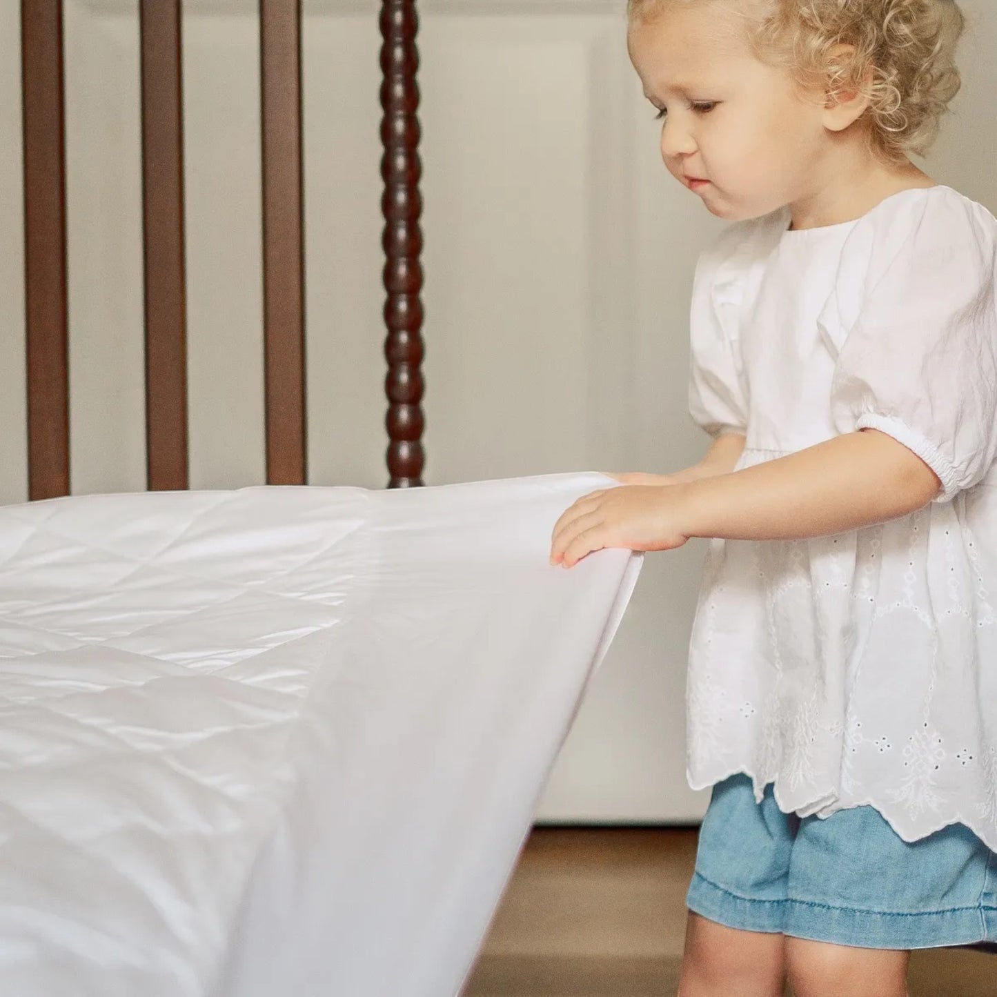A toddler in a white top touches a Modugreen Baby organic quilted white waterproof crib mattress protector pad.