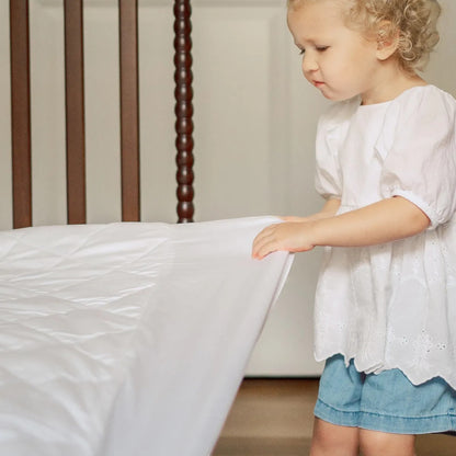 A toddler in a white top touches a Modugreen Baby organic quilted white waterproof crib mattress protector pad.